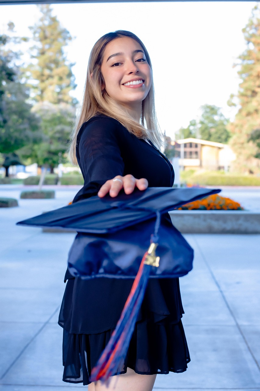 A smiling graduate holding a blue graduation cap toward the camera outdoors