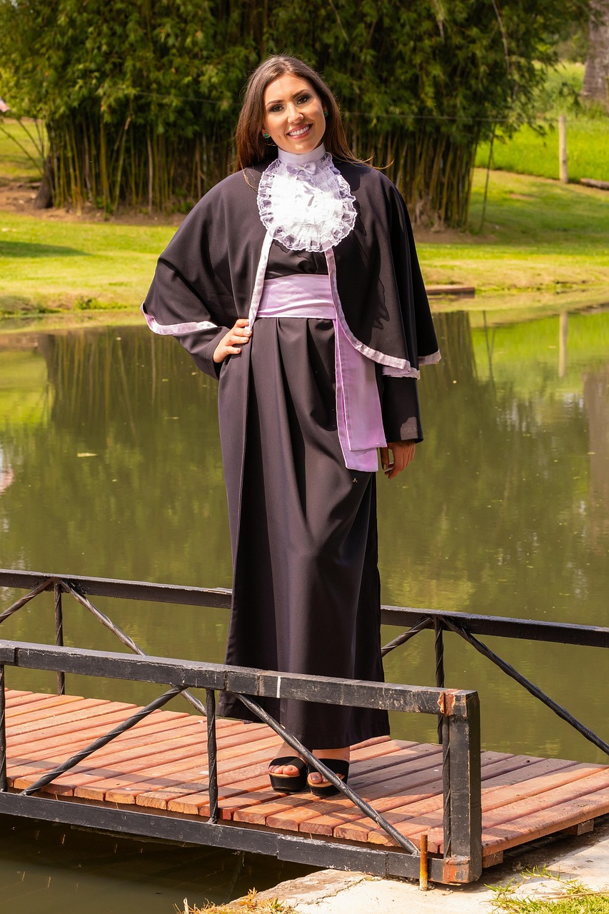 A graduate in a black gown standing on a small wooden bridge in a green park