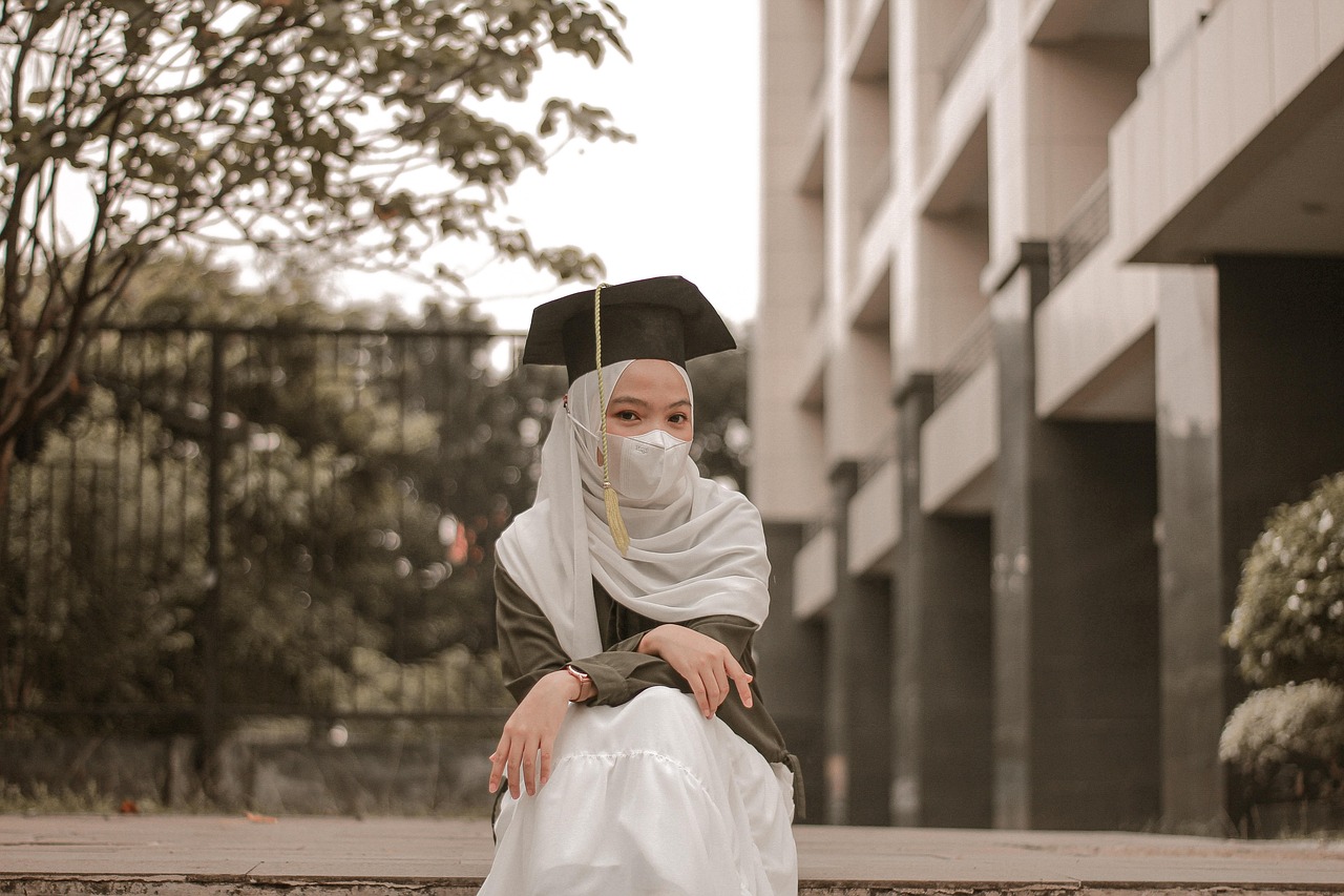 A graduate wearing a light-colored hijab and graduation cap seated outside a campus building