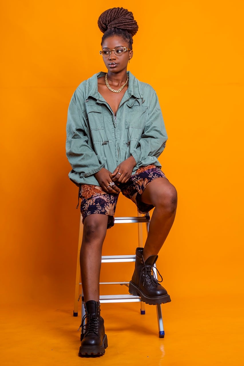 A woman with glasses and a braided updo seated on a stool against a bright orange backdrop