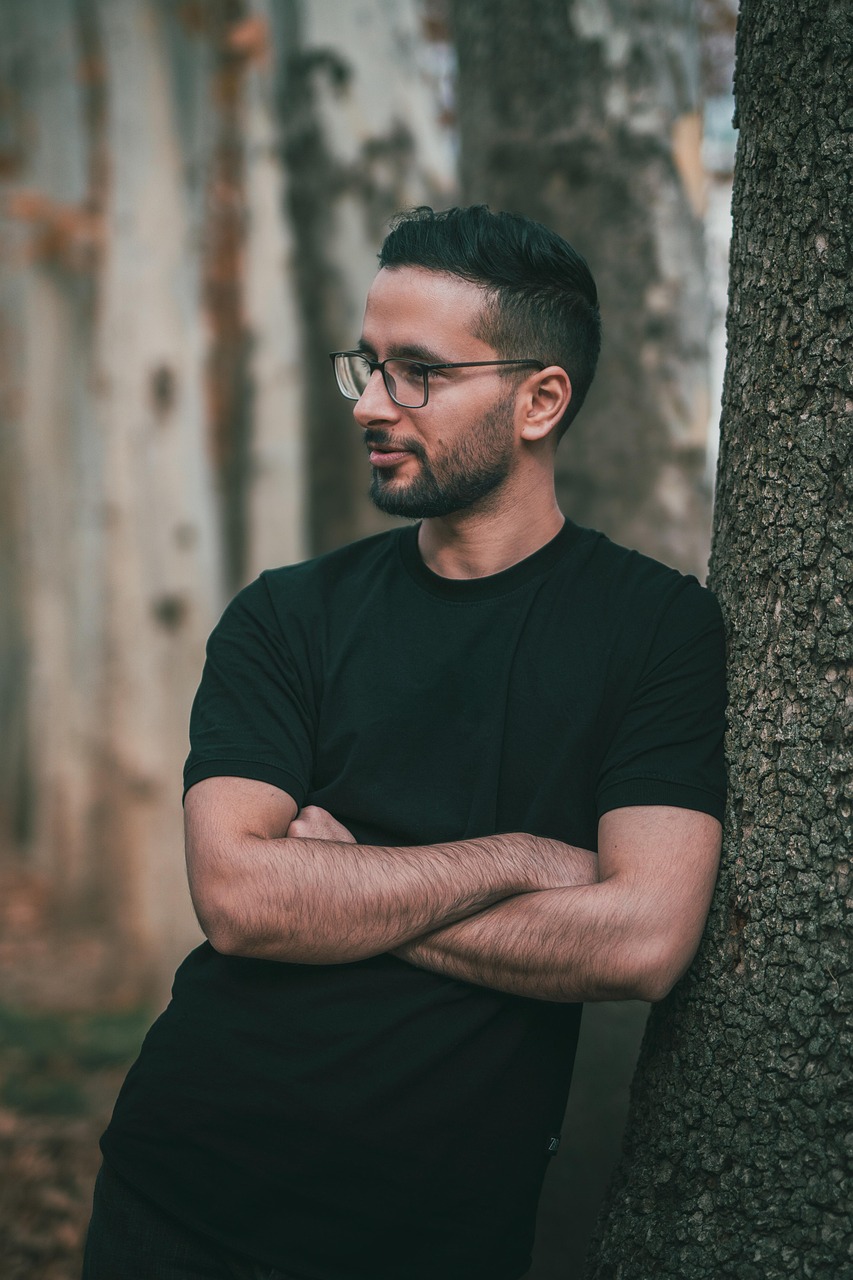 A man wearing a black shirt standing with folded arms beside a tree