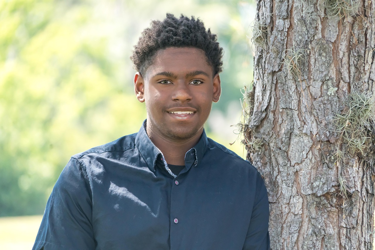 A young man in a blue button-up shirt standing beside a tree with a green background
